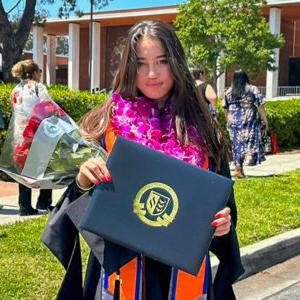 Madison Quan wears black graduation robe with a purple lei, a gray stole, and an orange and blue stole. Quan holds up the diploma from CGU and tucks a bouquet of red roses in her left arm. Quan has long straight dark brown hair.