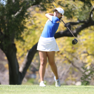 Katelyn Vo makes a stroke on the golf course. Vo wears the golf uniform of a white cap and white skirt with a blue shirt.